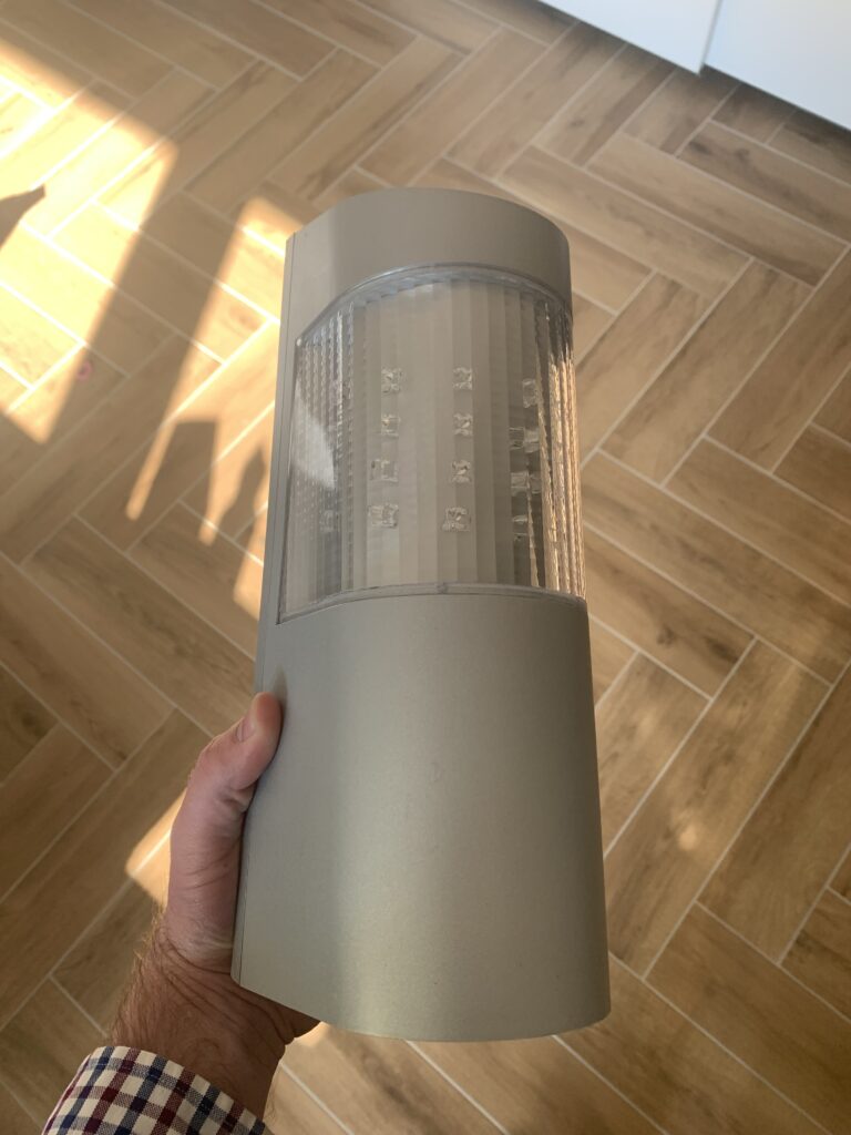 Eine Person hält eine moderne, graue Wandleuchte mit einem transparenten Einsatz für LEDs. Der Boden hat hellbraune Fliesen in Fischgrätmuster.