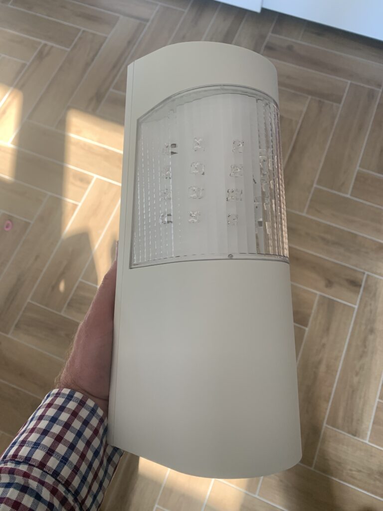 Ein Hand hält eine moderne Außenleuchte mit schlichtem, gebogenem Design und durchsichtigen Bereichen, durch die LED-Lichter sichtbar sind. Hintergrund zeigt einen Holzfußboden.