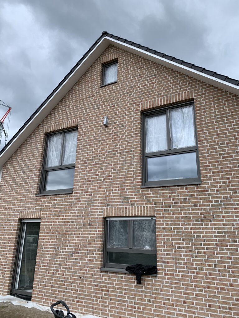 Ziegelhaus mit Satteldach und grauen Fenstern. Die Fenster sind mit Plastikfolie abgedeckt, was auf Renovierungs- oder Bauarbeiten hindeutet. Der Himmel ist wolkig.