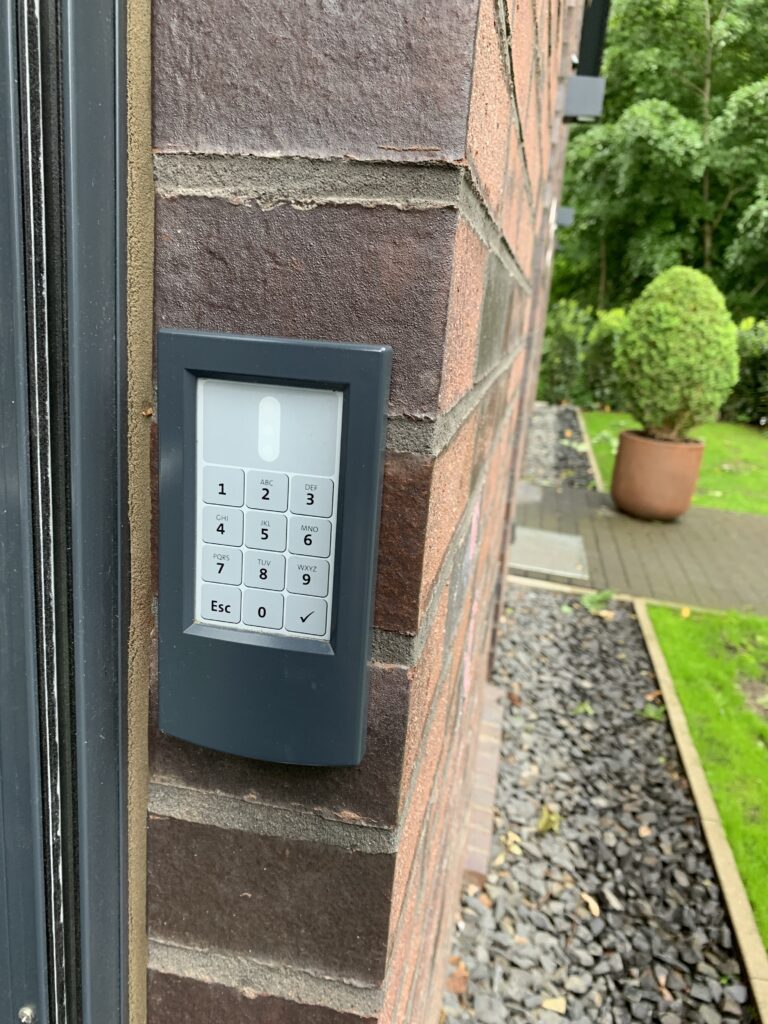 Ein elektronisches Tastenfeld mit einem Nummernblock ist an einer Backsteinwand montiert. Das Tastenfeld hat Tasten von 0 bis 9, sowie Esc und eine Bestätigungstaste.