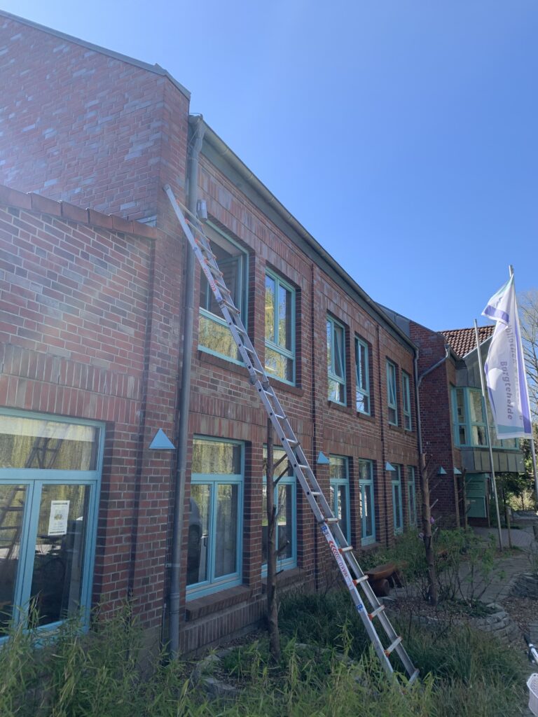 Eine lange Leiter lehnt an einem zweistöckigen roten Backsteingebäude mit blauen Fenstern. Sie reicht vom Boden bis zum Dach. Der Himmel ist klar und blau.