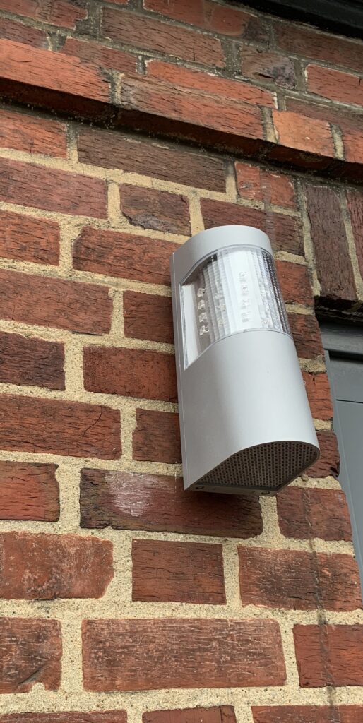 Eine moderne Außenleuchte aus Metall ist an einer Ziegelwand befestigt. Die Leuchte hat ein rundes Design mit einem transparenten oberen Bereich für die Lichtquelle.