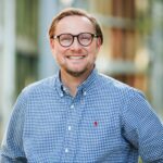 Ein lächelnder Mann mit Brille und Bart steht draußen. Er trägt ein blau-weiß kariertes Hemd. Im Hintergrund sind unscharf Gebäude und Bäume zu sehen.